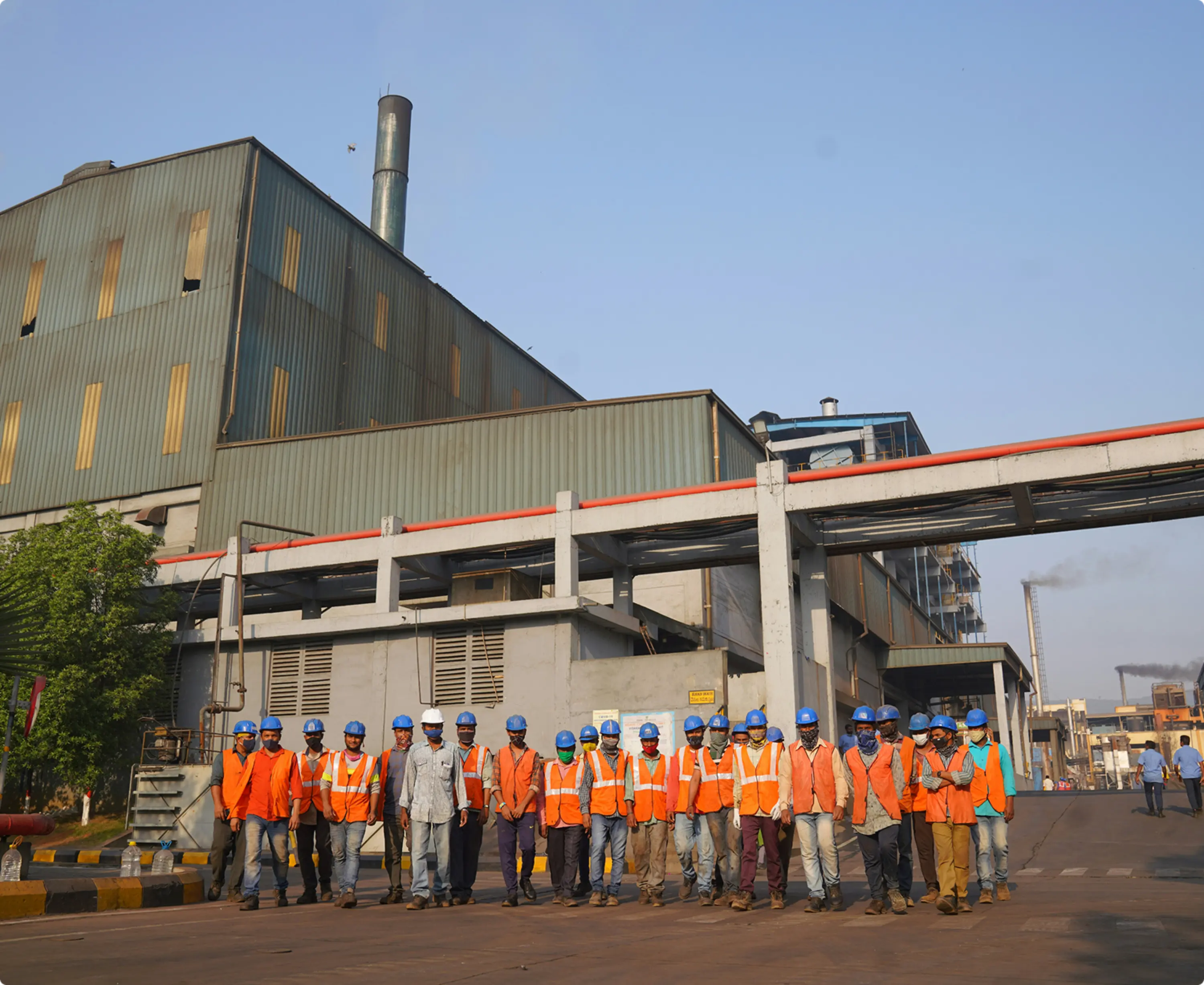 Un grupo grande de trabajadores con cascos y chalecos naranjas posando en una fábrica.